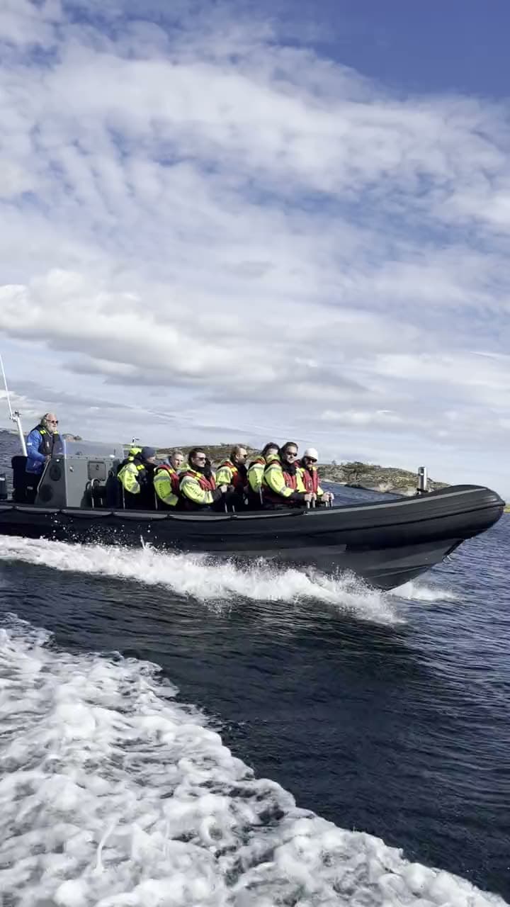 En RIB full av gjester i gule flytedresser i full fart forbi skjærene, fotografert fra båten ved siden av